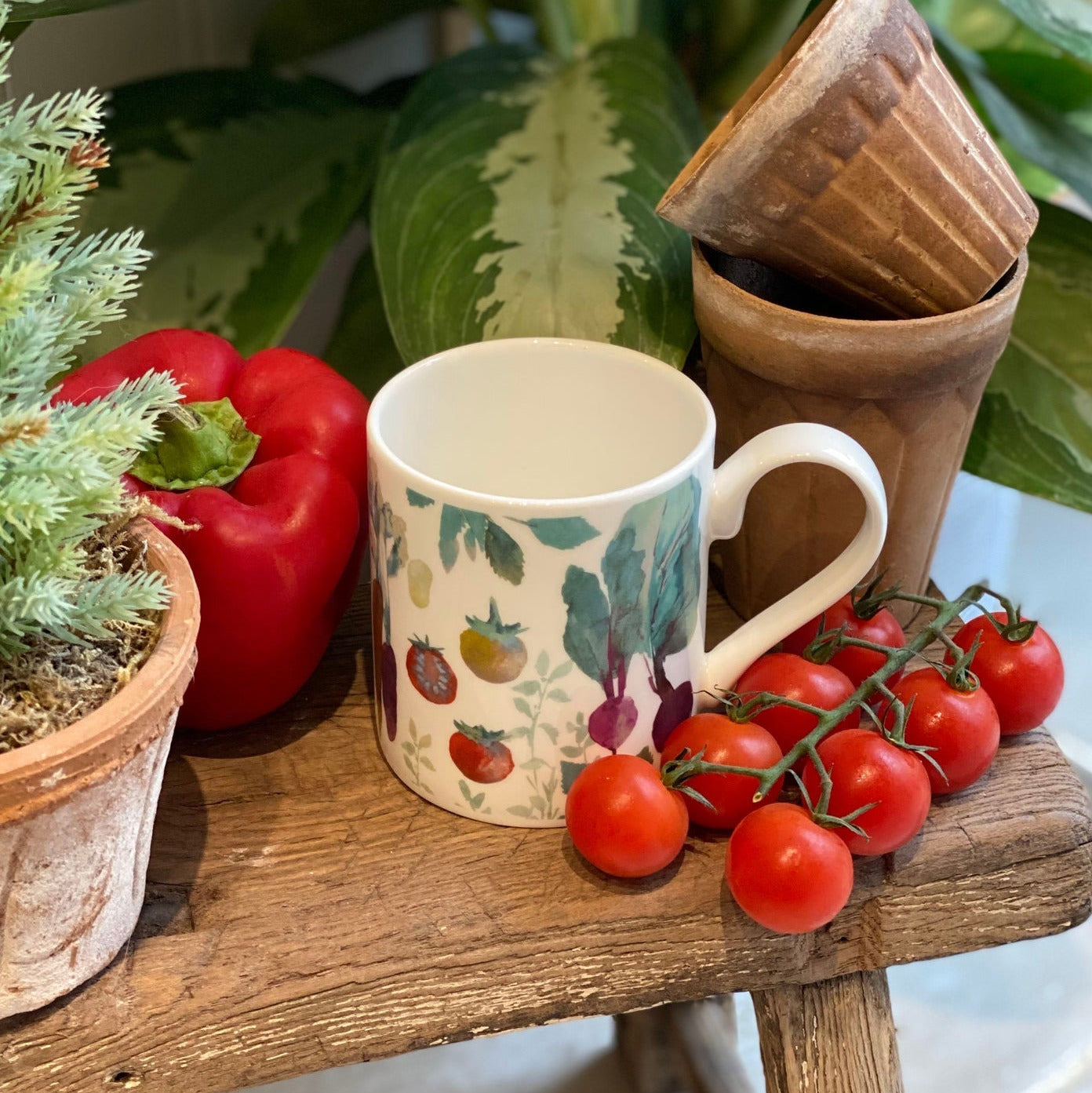 Mug with vegetable design on a wooden surface with tomatoes, bell pepper, and plants.
