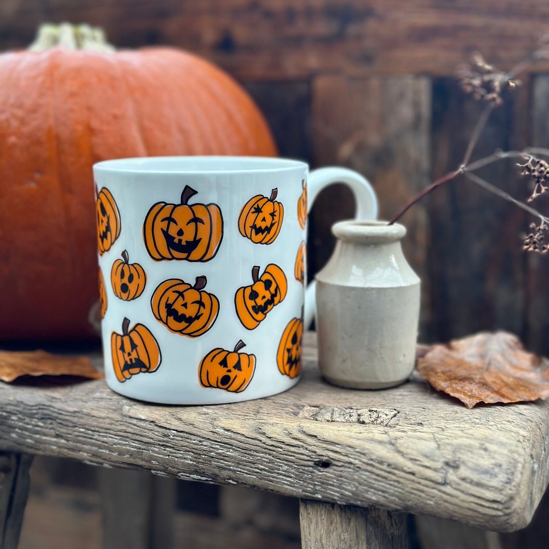 White mug with Halloween pumpkin design on a wooden surface with a pumpkin and small vase in the background.