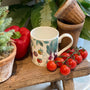 Mug with vegetable design on a wooden stool with tomatoes and bell pepper, surrounded by plants.