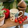 Mug with vegetable design on a wooden surface with tomatoes, bell pepper, and plants.