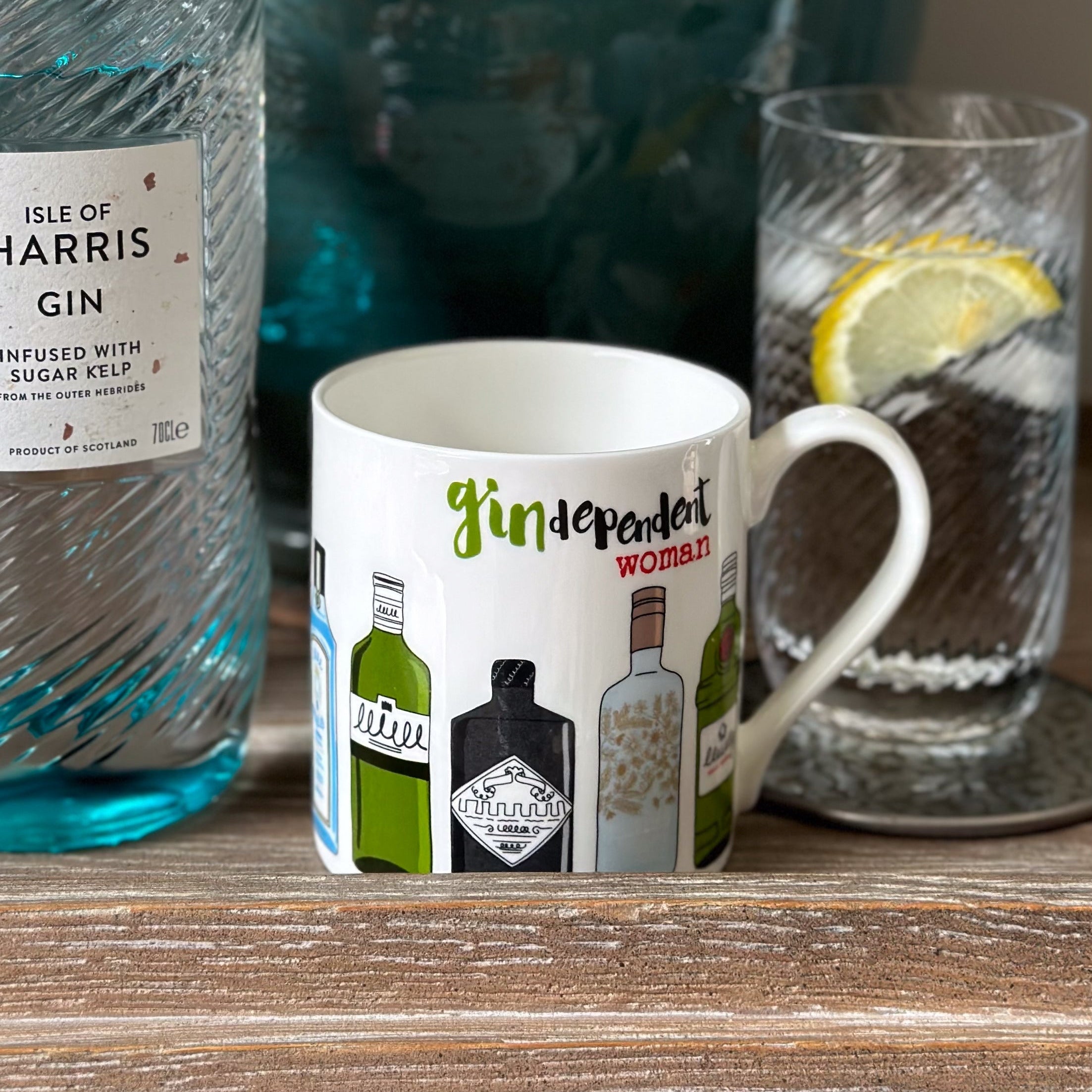 Gin bottle, mug with gin-themed design, and glass with lemon slice on a wooden surface.