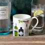 Gin bottle, mug with gin-themed design, and glass with lemon slice on a wooden surface.