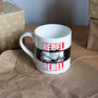 White mug with 'REBEL REBEL' design on a brown surface with wrapped gifts in the background