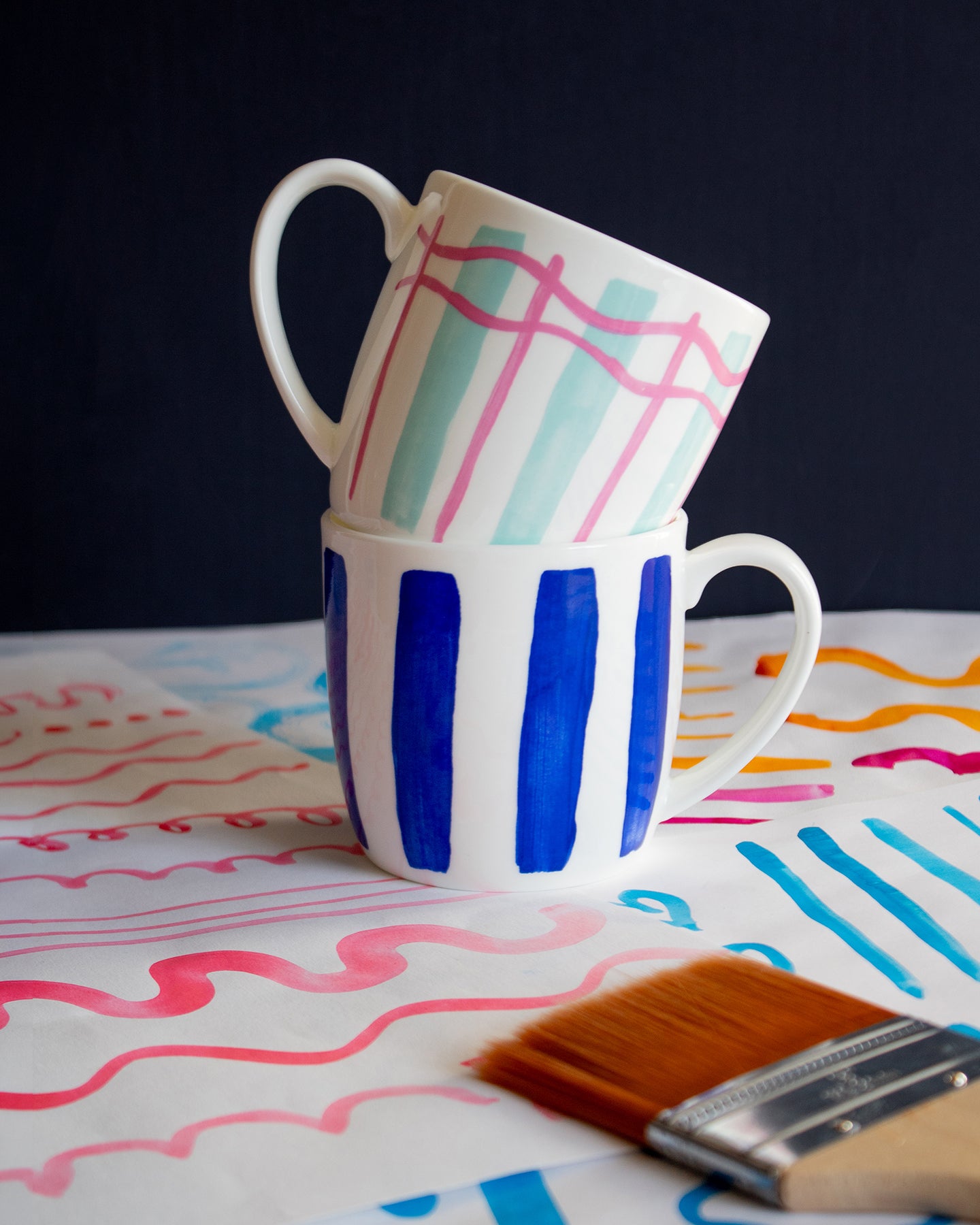 Two mugs with plaid and striped patterns stacked on a table with paintbrush and colorful paint strokes.
