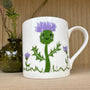 Mug with a cartoon thistle design on a wooden surface with a thistle plant in the background.