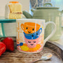 Colorful mug with cartoon-style teacups on a wooden surface with strawberries and a cake in the background.