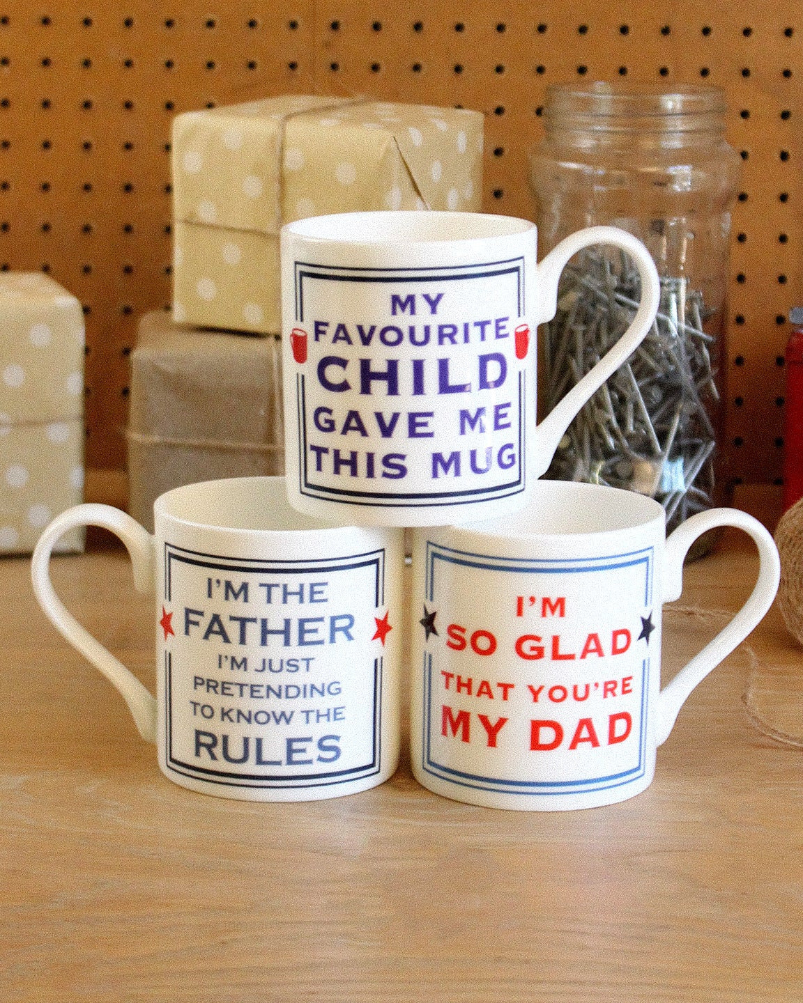 Three mugs with humorous text on a wooden surface with a pegboard background.