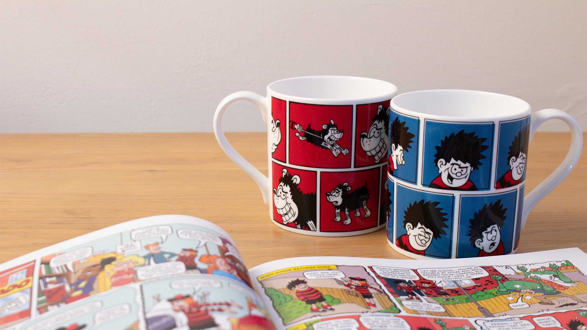 Two mugs with cartoon character design on a wooden surface next to an open book.
